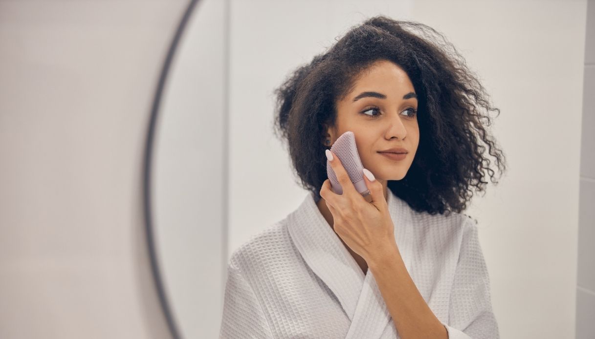 ragazza mulatta capelli afro con accappatoio bianco pulisce il viso con una spazzola davanti allo specchio