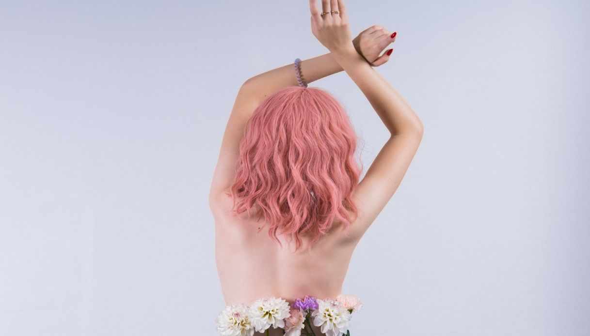 ragazza di spalle con capelli rosa pastello mossi, schiena nuda, mani in alto e fiori nei pantaloni