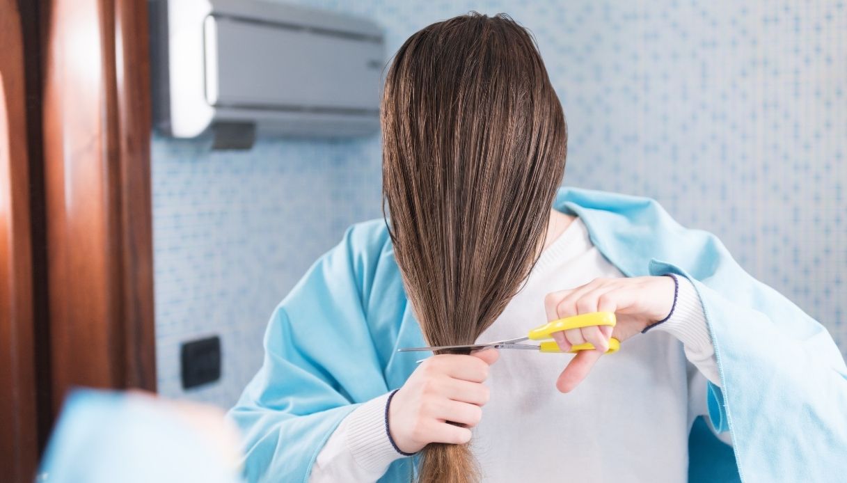 ragazza con capelli lunghi davanti la faccia li taglia con forbici gialle e un asciugamano azzurro sulle spalle