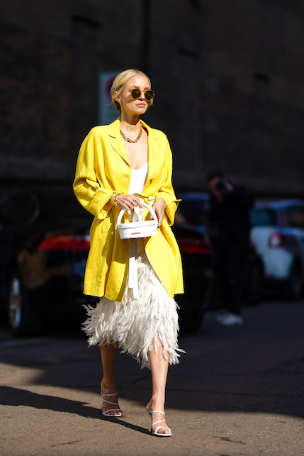 modella con giacca gialla, vestito bianco con piume, tacchi e collana dorata