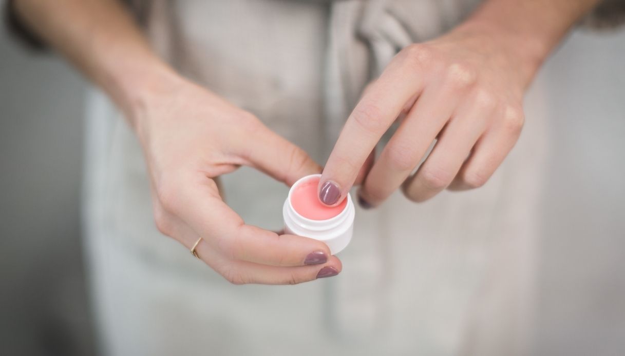 mani femminili con smalto violaceo toccano balsamo labbra rosa in barattolo bianco