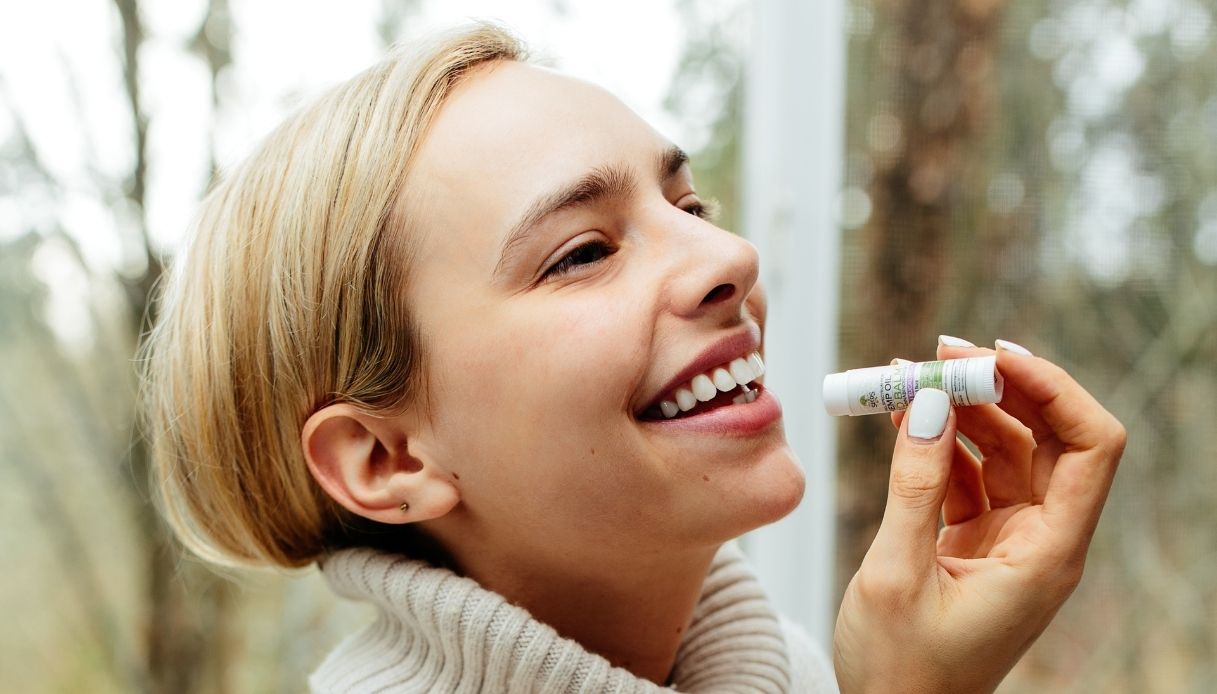 profilo di donna bionda con capelli dentro il maglione che sorride e sta per applicare balsamo labbra con mani curate e unghie bianche